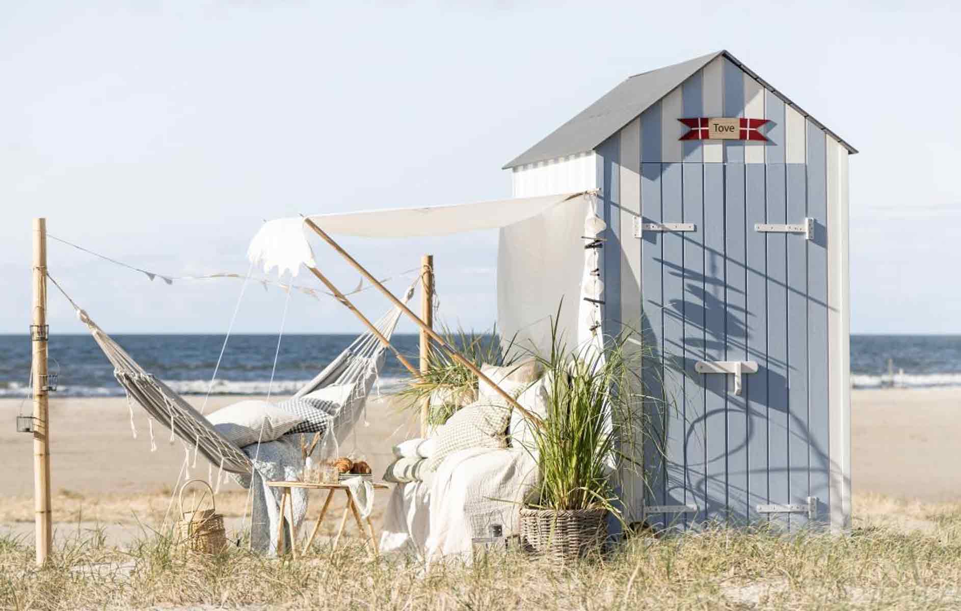 Eine ruhige Strandkulisse mit einem kleinen Strandhaus mit der Aufschrift „Tiki-Bar“, einer Hängematte und gemütlichen Sitzgelegenheiten, umgeben von Topfpflanzen und einem weißen Stoffbehang.