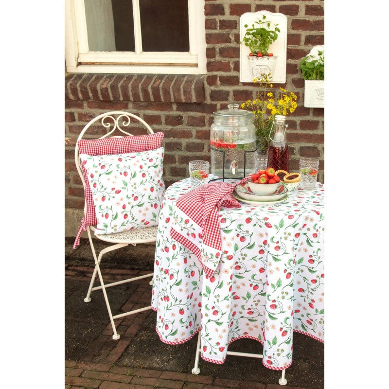 Gartentisch mit Tischdecke mit Kirschmuster, einer Glaskanne, zwei Gläsern, einem Teller mit Erdbeeren und einem weißen Stuhl mit rotkariertem Kissen vor einer Backsteinwand mit hängenden Blumentöpfen.