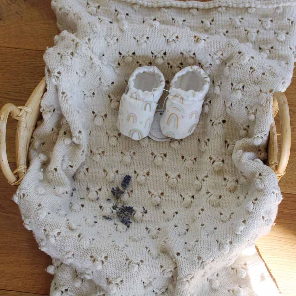 Weiße Babydecke mit einem kleinen Lavendelstrauß und Cosy Roots - Lauflernschuhe-Regenbögen darauf, alles in einem Holzkorb ruhend.