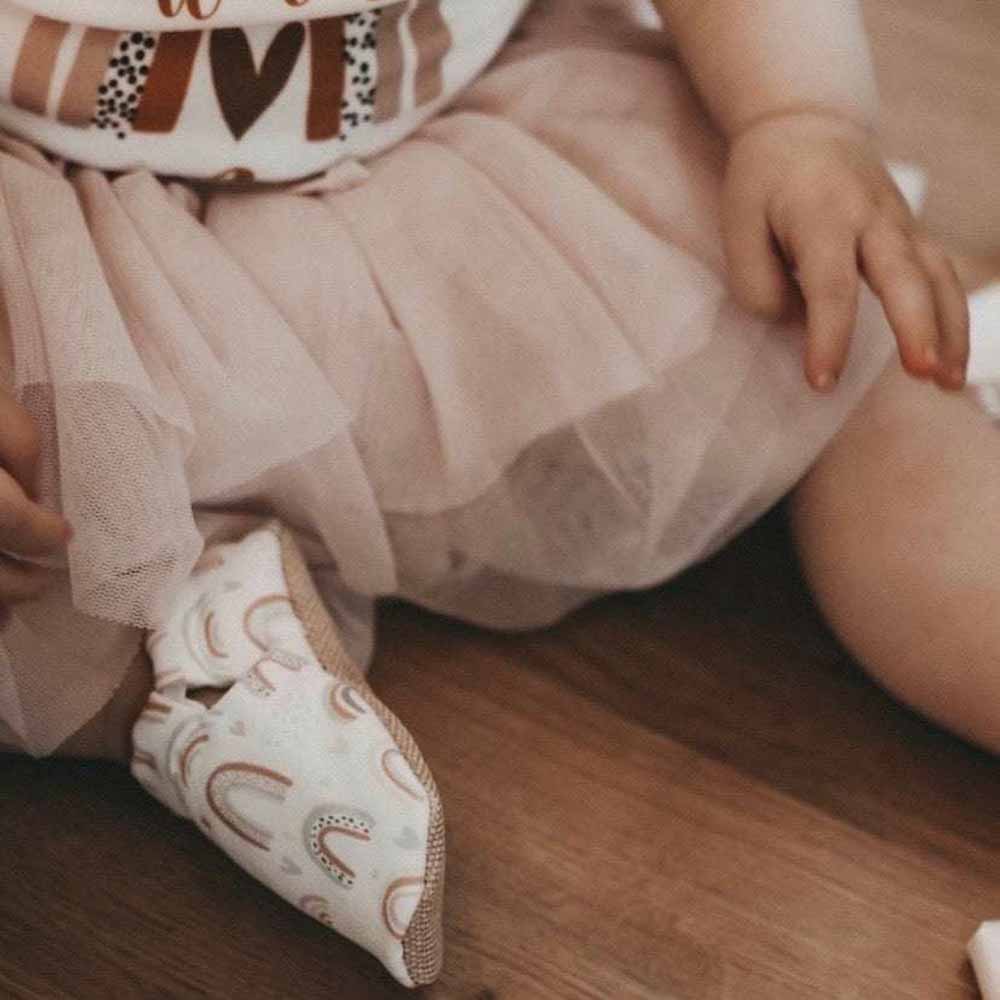 Ein Baby trägt Lauflernschuhe „Regenbogen“ von Cosy Roots und einen rosa Tüllrock, wobei der Schwerpunkt auf der Textur des Stoffes und den gemusterten Socken liegt.
