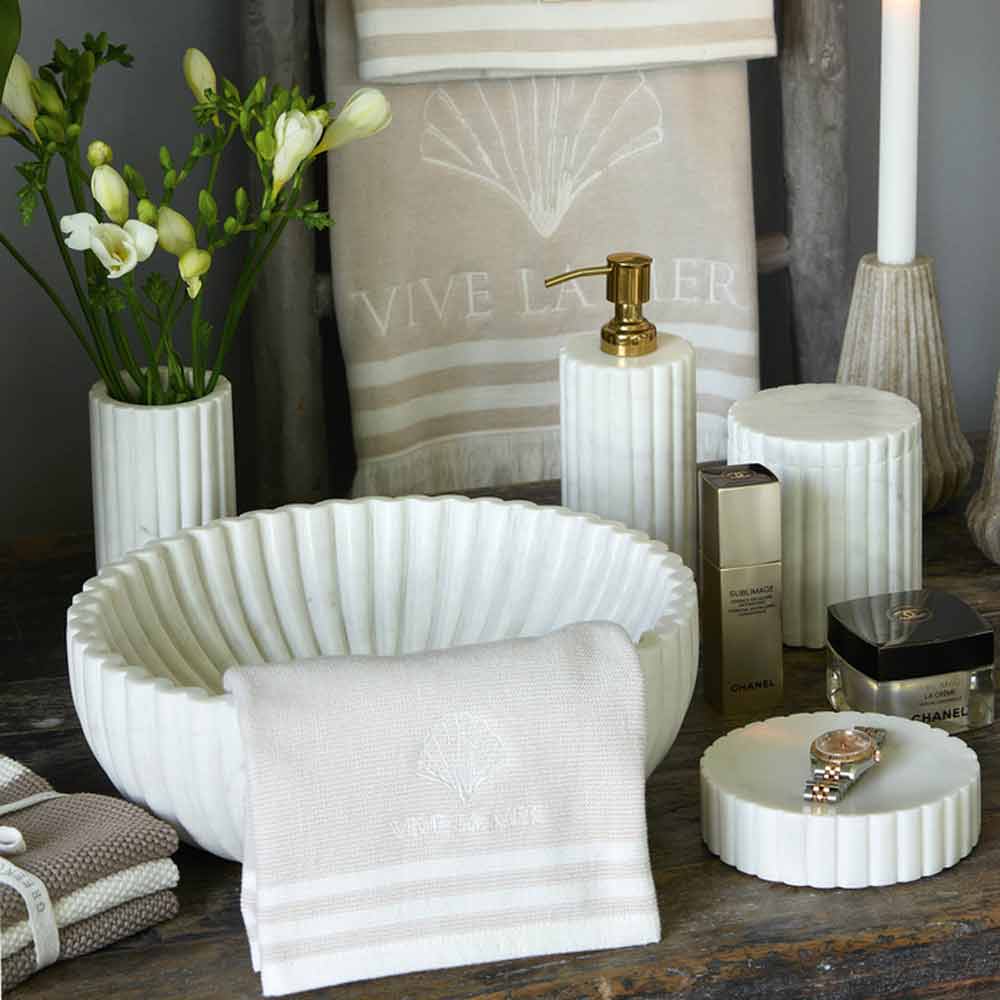 Elegante Badezimmeraccessoires, darunter Handtücher, Seifenspender, Kerzen und GreenGate Marmor Gefäß mit Deckel auf einem grauen Tisch.