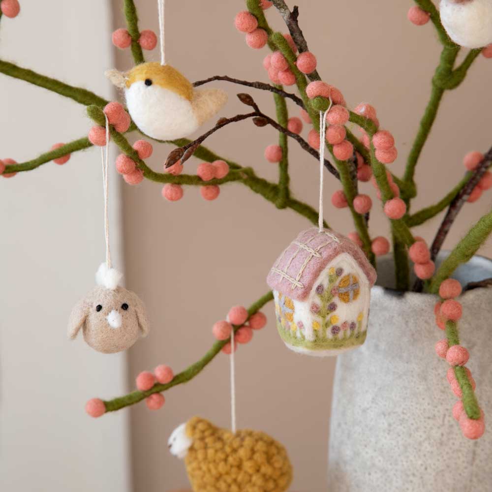 An den Zweigen hängende Filzkunstwerke, darunter ein Anhänger Haus aus Filz von Gry & Sif, ein Vogel und ein Häschen, präsentiert in einer Vase mit rosa akzentuierten Zweigen.
