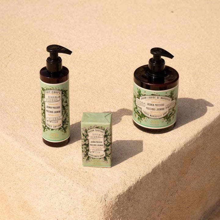 Zwei 150-g-Pumpflaschen „Panier des Sens Feste Seife mit Duft Jasmin“ und ein kleines Sachet mit grünen Etiketten, platziert auf einer Betonfläche im Sonnenlicht.