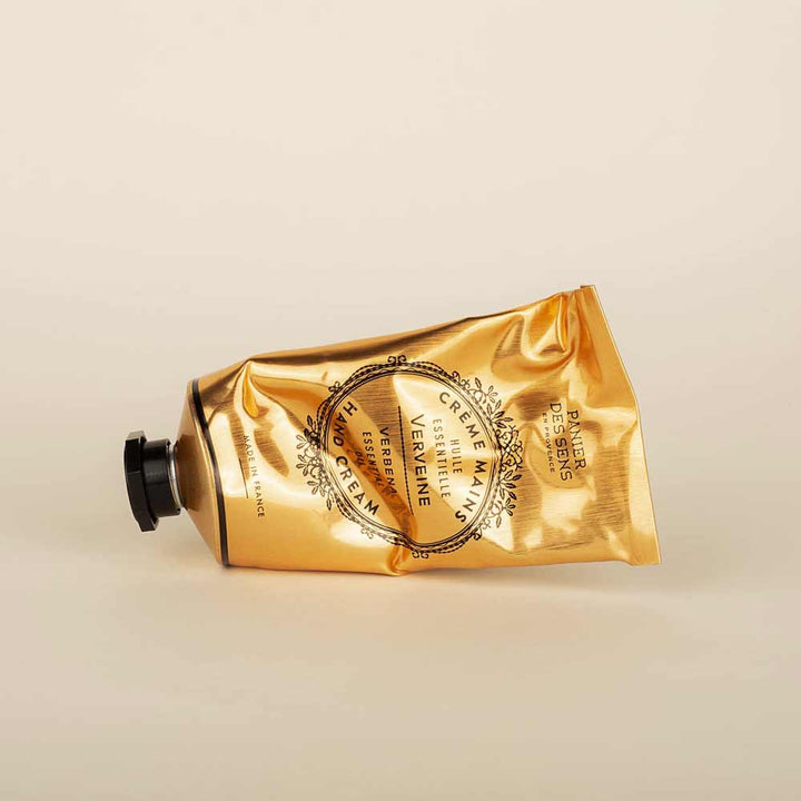 Eine goldene Tube Panier des Sens – Duschgel Eisenkraut (Verbene) 250 ml, die auf der Seite liegt, vor einem neutralen beigen Hintergrund, beschriftet mit elegantem schwarz-weißem Text.