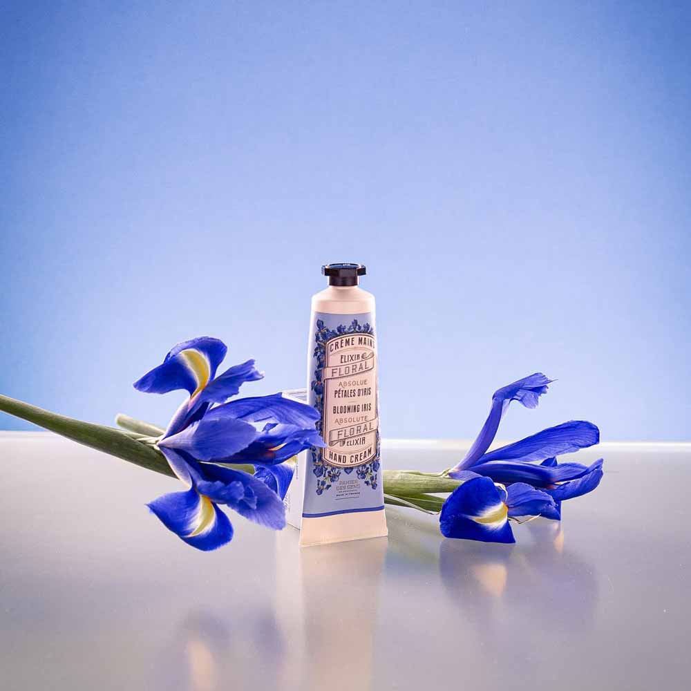 Eine Flasche Panier des Sens - Handcreme Irisblüttenblätter neben frischen blauen Irisblüten auf einer reflektierenden Oberfläche mit blauem Hintergrund mit Farbverlauf.