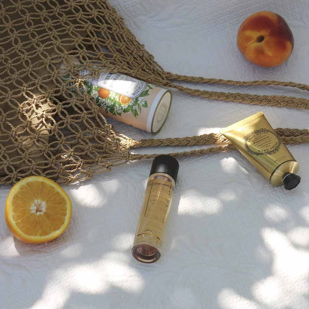 Auf einer weißen Decke angeordnete Sommer-Essentials, darunter eine Panier des Sens – Handcreme Provence, Sonnencreme, Pfirsich und eine Orangenscheibe.