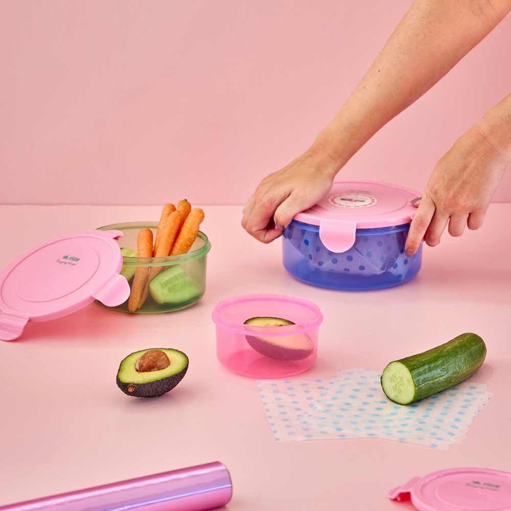 Hände organisieren Reis – Lunchbox rund, mehrfarbig, 3er-Set, Behälter mit Obst und Gemüse auf einem rosa Tisch.