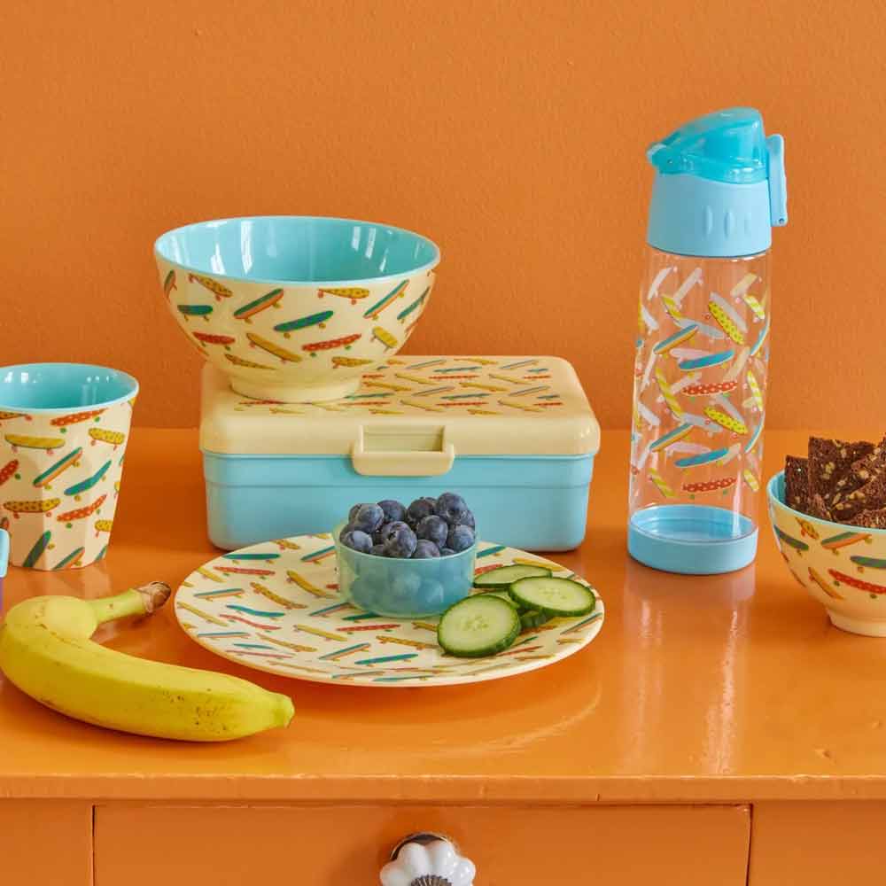 Eine Auswahl an Mittagsgerichten auf einem orangefarbenen Tisch, darunter ein gemusterter Teller mit Obst und Gurken, eine Sandwichbox, ein Rice - Melaminbecher-Skateboard mittlerer Größe, eine Wasserflasche und Snacks.
