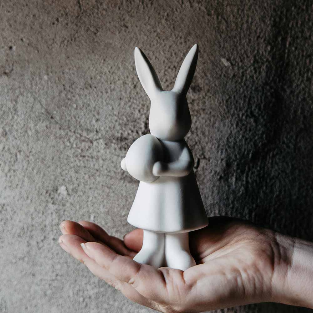 Eine Hand hält eine weiße Kaninchenfigur von Storefactory - Alice Hase, die ein kleineres Kaninchen vor einem strukturierten grauen Hintergrund hält.