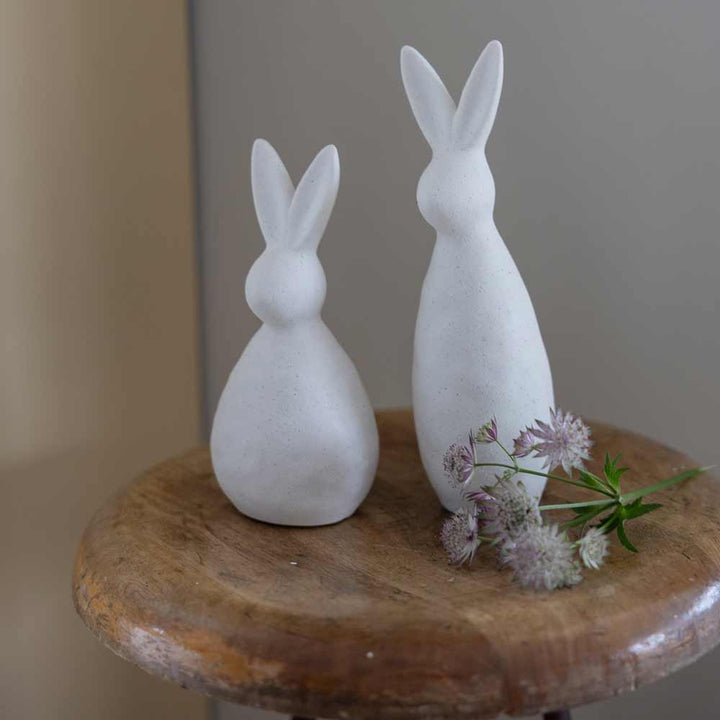 Zwei Storefactory - Alma Hase Figuren auf einem Holzständer mit einem Blumenzweig.