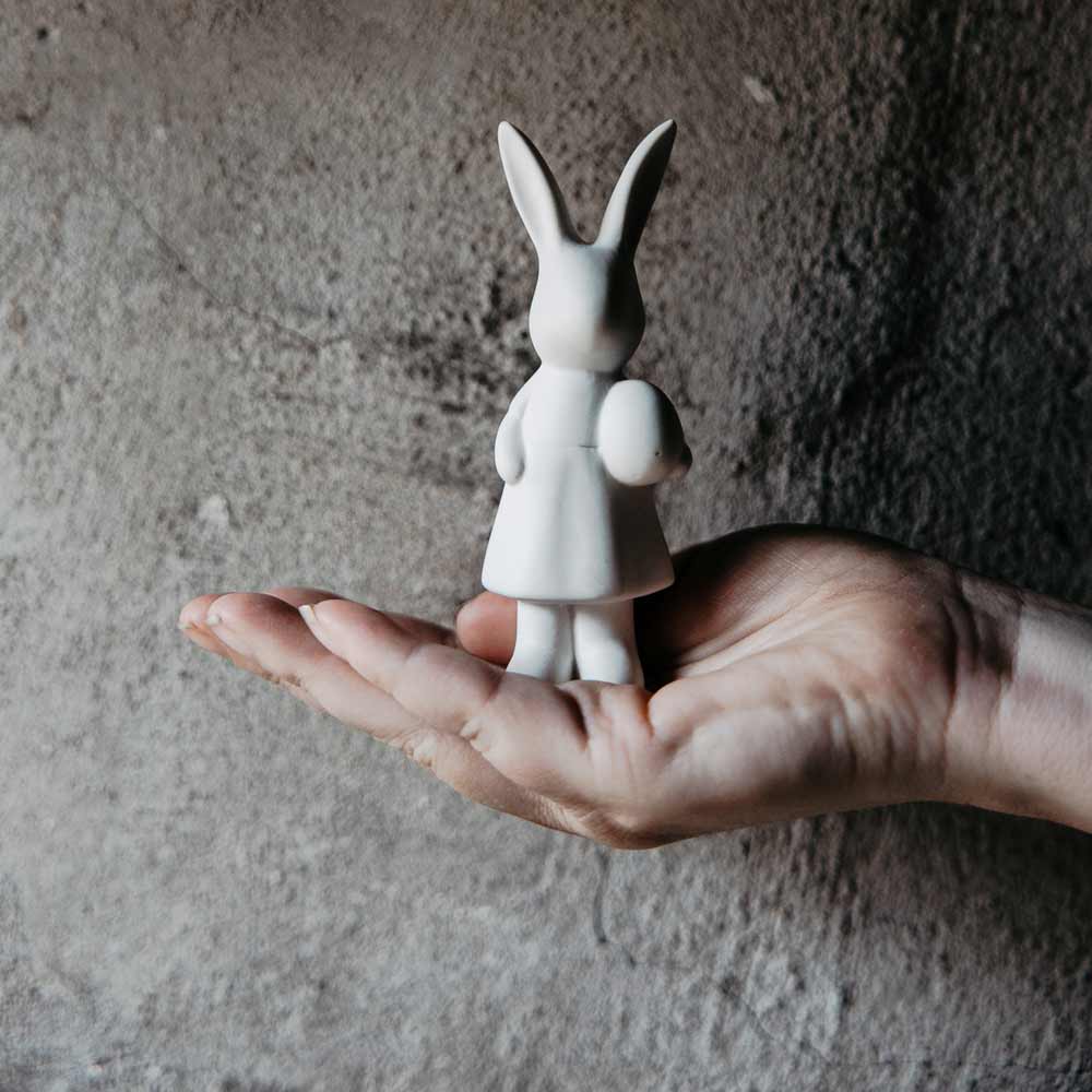 Eine Storefactory - Ester Hase-Figur eines Kaninchens, das ein Kind hält, liegt in der Handfläche vor einem strukturierten grauen Hintergrund.