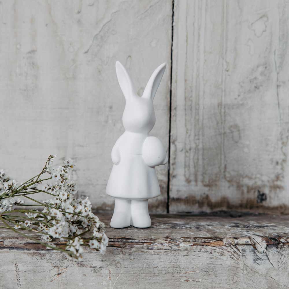 Eine weiße Storefactory - Ester Hase-Figur eines Hasen, der neben kleinen weißen Blumen auf einer rustikalen Holzoberfläche vor einem verzweifelten Hintergrund steht.