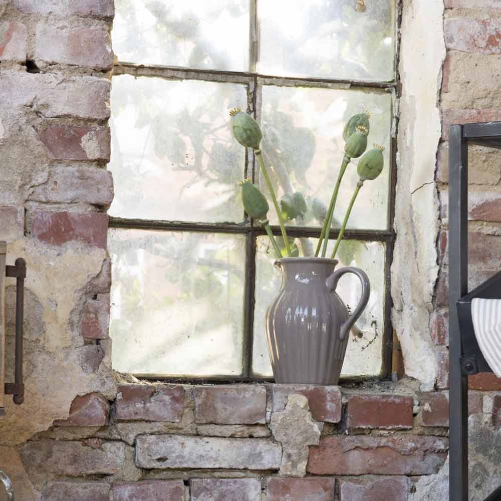 Ein Ib Laursen - Krug Mynte 1,7 Liter Keramikkrug mit mehreren Kaktusstängeln steht auf einem gemauerten Fensterbrett vor einem alten, rustikalen Fenster.