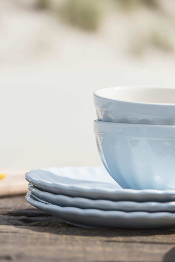 Stapel weißer Ib Laursen - Müslischale Mynte-Teller und eine Teetasse auf einem Holztisch vor einem verschwommenen Sandstrand-Hintergrund.