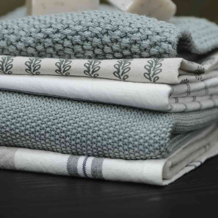 Ein Stapel ordentlich gefalteter Textilien, darunter eine gehäkelte graue Decke und Handtücher vom Typ Topflappen Mynte von Ib Laursen.
