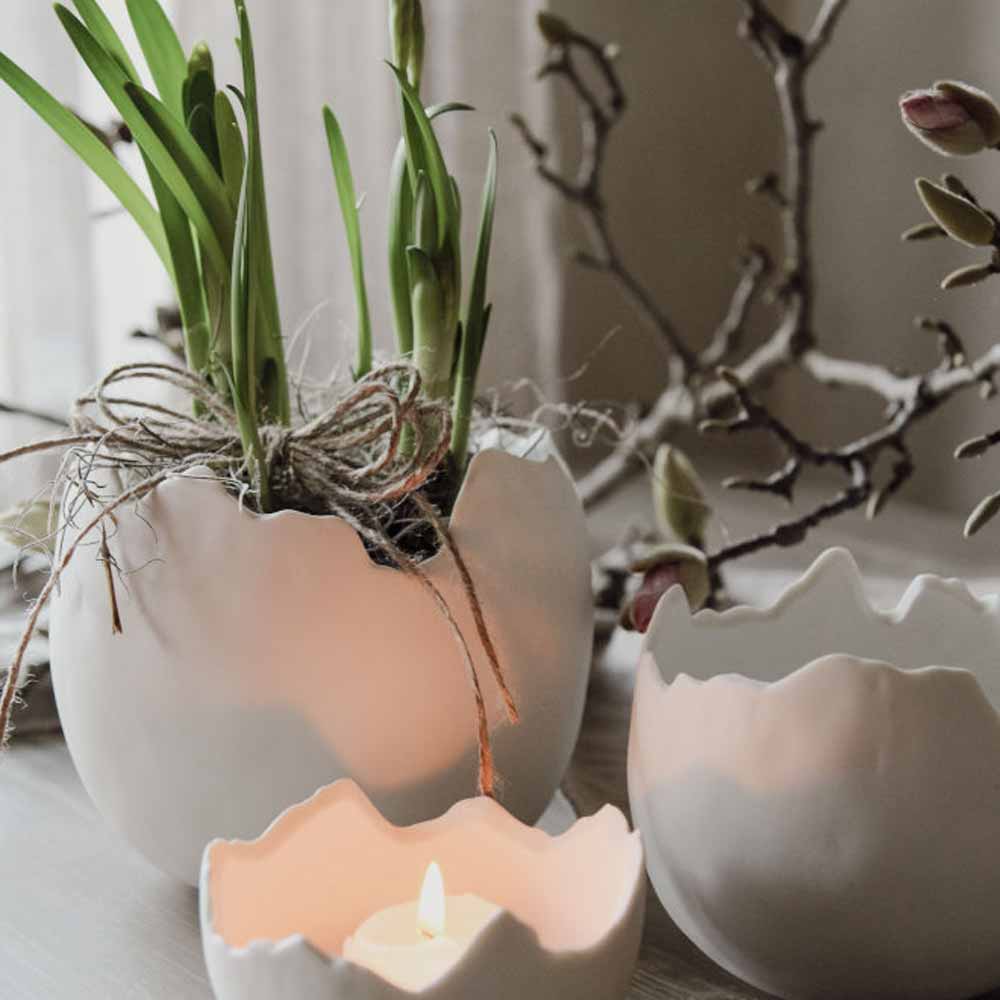 Pflanzen wachsen im Majas Cottage – Eierschale-Keramik-Pflanzgefäße mit einer brennenden Kerze daneben auf einem Tisch.