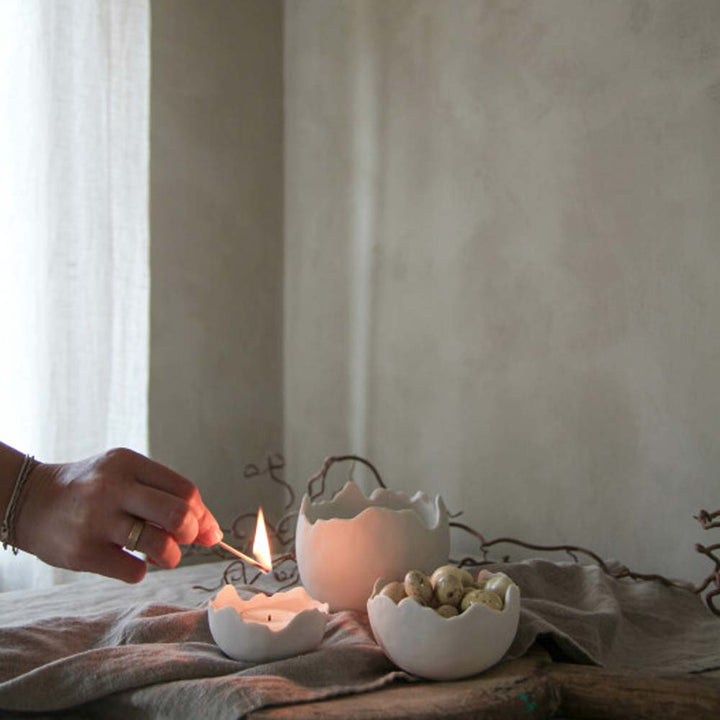 Eine Person zündet eine Kerze an, die in einer rissigen Majas Cottage - Eierschale Keramik platziert ist, während andere Majas Cottage - Eierschale Keramik mit kleinen Dekorationsgegenständen auf einer mit Stoff bedeckten Oberfläche liegt.