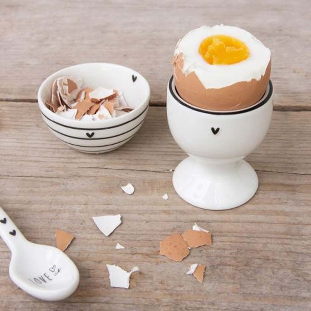 Weichgekochtes Ei in einer Bastion Collections – Eierbecher mit Fuß Little Heart schwarz mit verstreuten Eierschalenstücken auf einer Holzoberfläche.