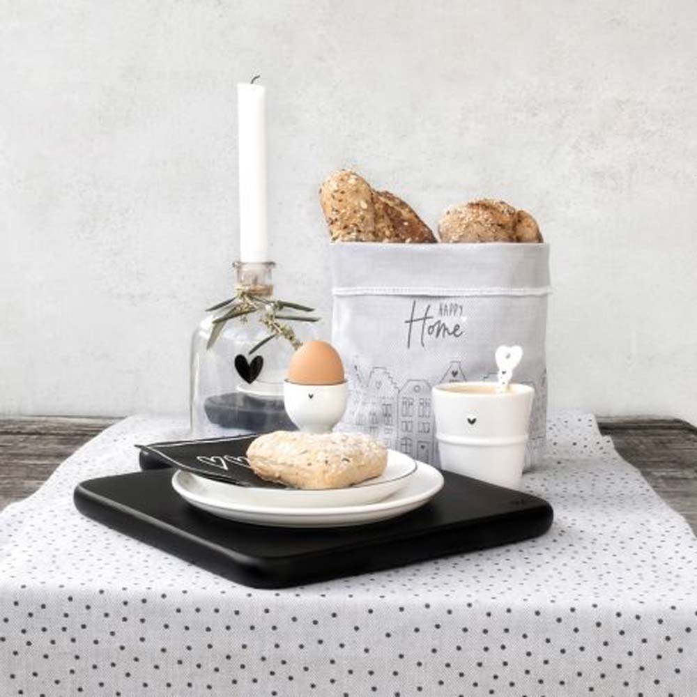 Ein gedeckter Frühstückstisch mit einem Bastion Collections – Eierbecher mit Fuß Little Heart schwarz, einem Brötchen auf einem schwarzen Teller, einer Kerze und einem dekorativen Stoffbeutel mit der Aufschrift „Home“.