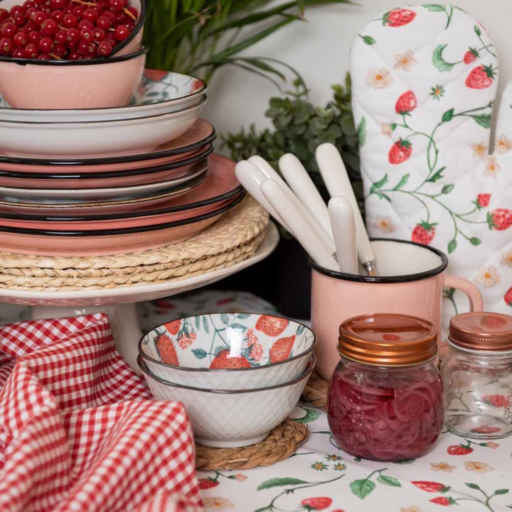 Satz mit Produktname: Tischgedeck mit gestapelten Tellern, einer Tasse mit Besteck, Einmachgläsern, einer Schüssel und einem Ofenhandschuh „Erdbeere“ von Clayre & Eef, alles zwischen grünem Laub arrangiert.