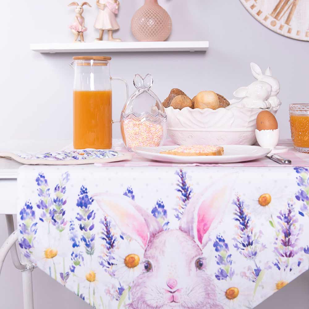 Ein mit Clayre & Eef – Vorratsdose Osterei mit Deckel gedeckter Frühstückstisch mit Tischdecke mit Hasenaufdruck, einem Krug Orangensaft und Schüsseln mit Eiern.
