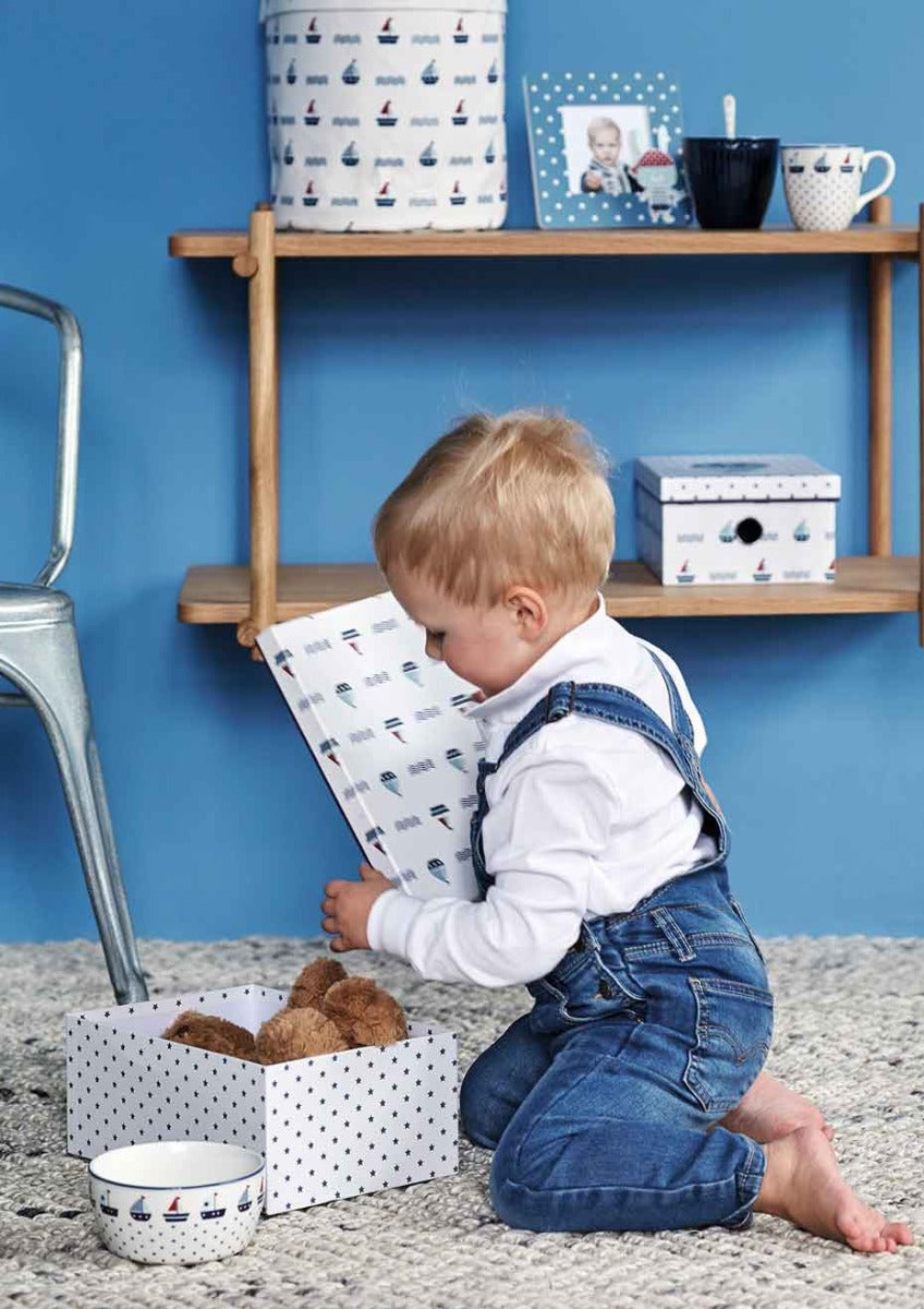 Kleinkind im Jeans-Overall, das ein Buch betrachtet und auf einem Teppichboden neben einer offenen Schachtel mit Teddybären in einem Raum mit blauen Wänden und Holzregalen sitzt.