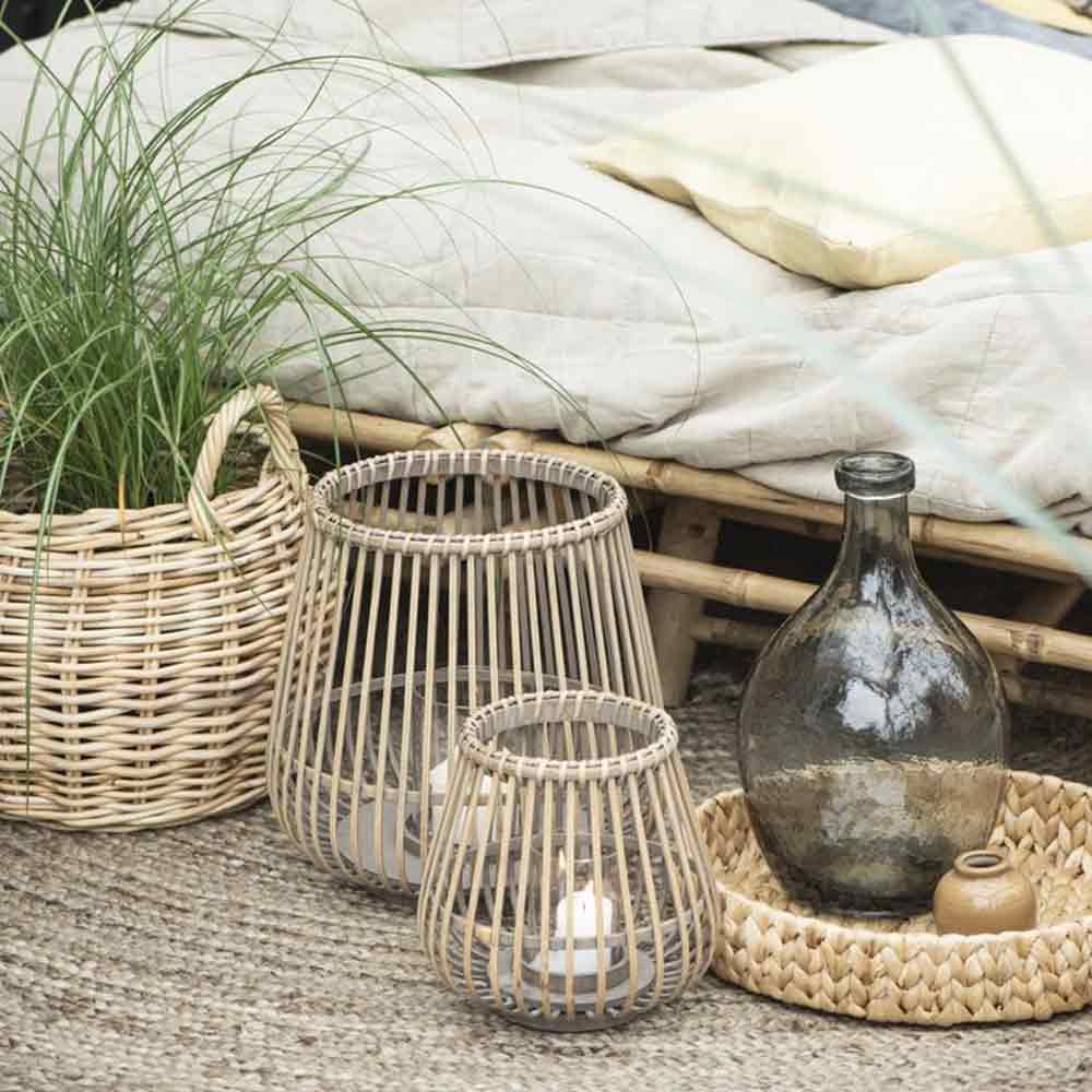 Runder Rattankorb mit Gras links, zwei offene Laternen und dunkle Glasflasche vor einer Sitzbank mit hellen Polstern.