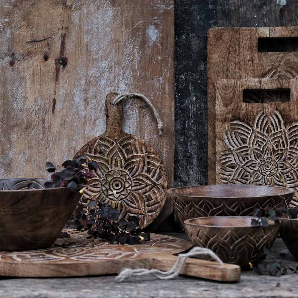Rustikales Küchengeschirr aus Holz von Majas Cottage mit aufwendigen Schnitzereien auf einem verwitterten Tisch, darunter Schüsseln, Schneidebretter und ein mit einem Blumenmuster verzierter Dekorationsgegenstand.