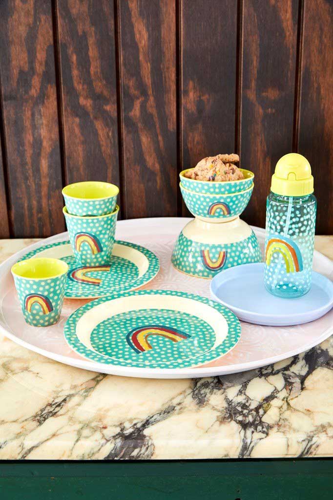 Farbenfrohes Picknick-Set bestehend aus Tellern, Tassen und Schüsseln mit Regenbogenmotiven, arrangiert auf einem Marmortisch vor einem Holzhintergrund. (Rice - Melaminbecher Rainbow & Stars Medium)