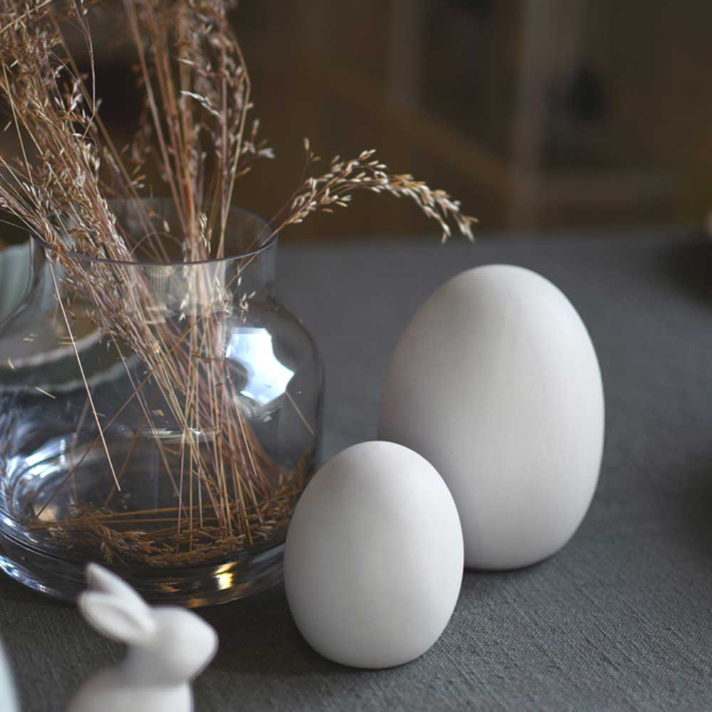 Zwei weiße Storefactory - Bjuv Ei Keramik unterschiedlicher Größe auf einer grauen Oberfläche mit einer Glasvase mit getrockneten Pflanzen und einer kleinen weißen Kaninchenfigur.
