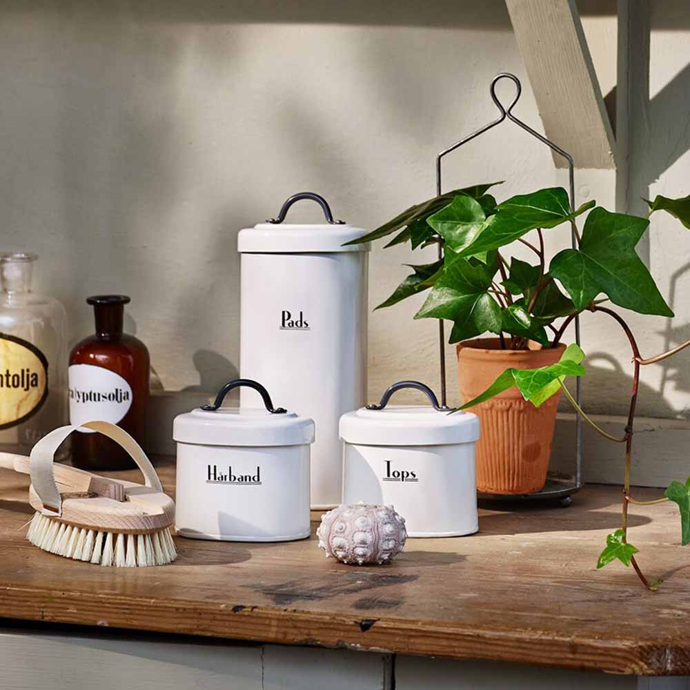 Weiße Strömshaga – Vorratsdose. Deckel mit der Aufschrift „Pasta“, „Havregry“ und „Løps“ und schwarzem Deckel, auf einem Holzregal neben einer Pflanze und einer Bürste.