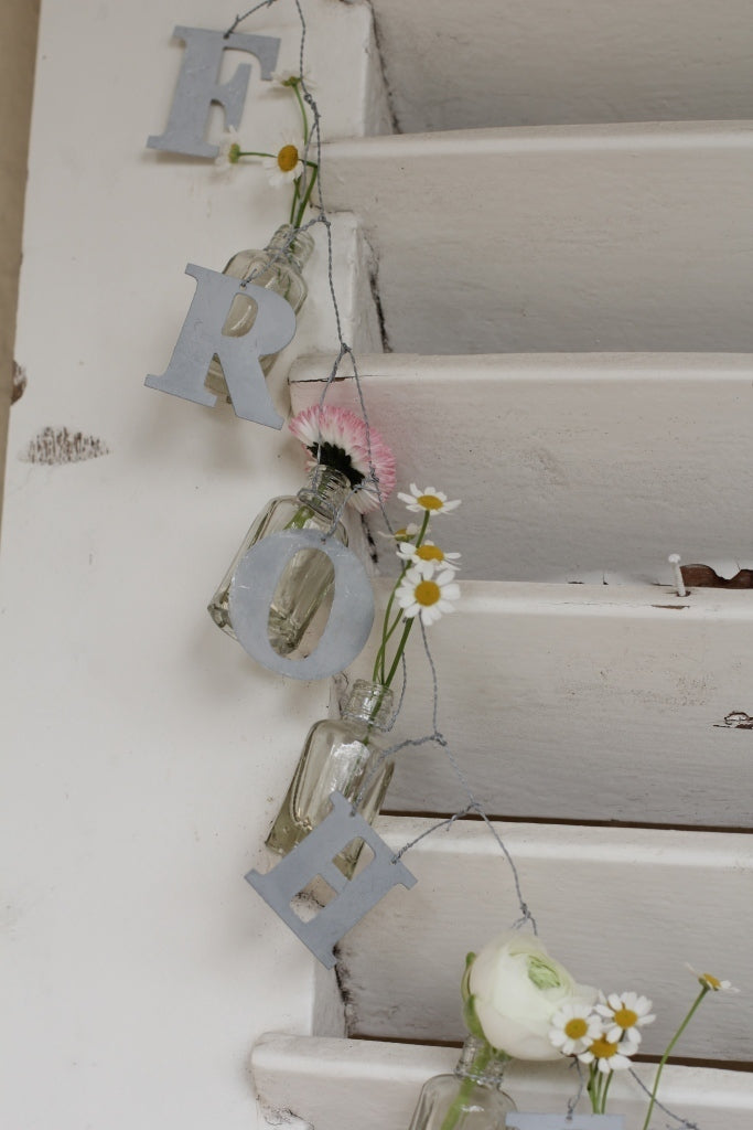 Dekorative Buchstaben mit der Aufschrift „Vl Home – Girlande Frohe Ostern“ mit kleinen Blumen in Klarglasvasen, die an einer weißen Treppe hängen.