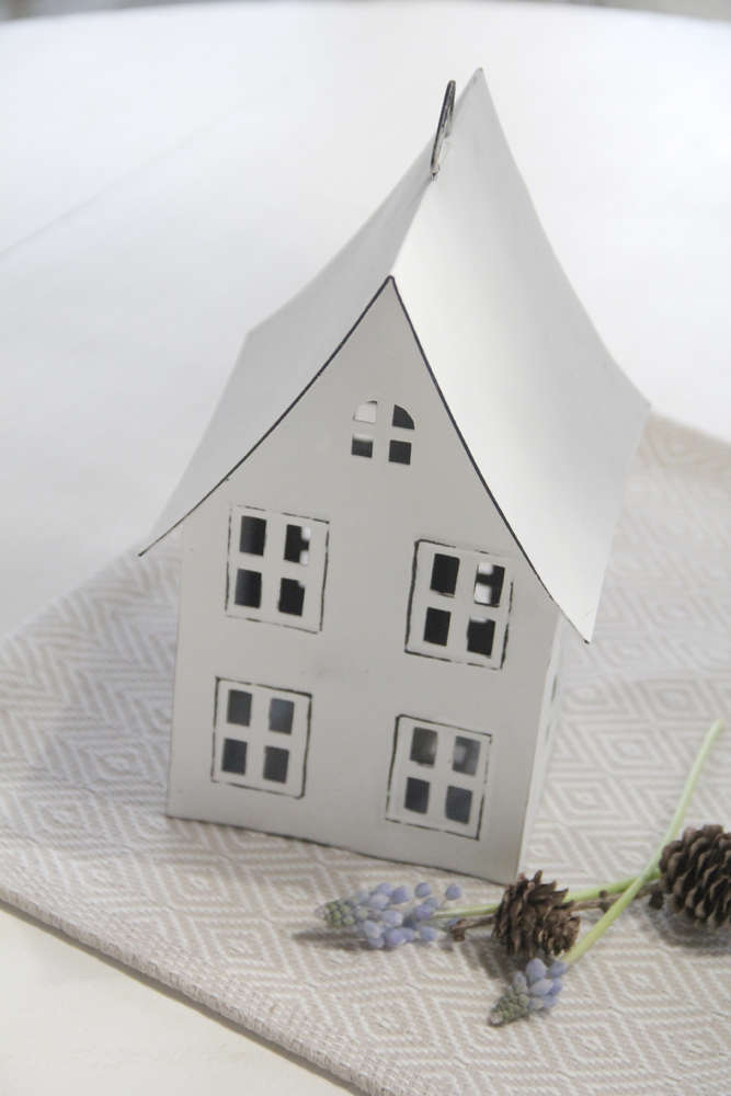 Ein kleines, handgefertigtes Vl Home - Metallhaus weiß mit spitzem Dach-Modell mit geprägten Fenstern und einem spitzen Dach, platziert auf einem strukturierten weißen Tuch, begleitet von Tannenzapfen und getrockneten Blumen.