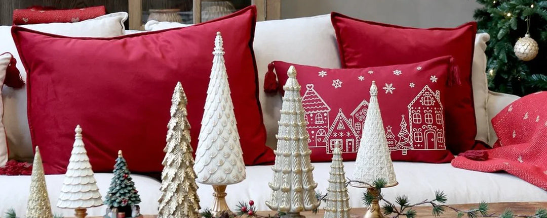 Ein rotes Zierkissen mit weißen Strichzeichnungen von Häusern und Schneeflocken, die an den Ecken mit Quasten versehen sind, liegt vor anderen roten und weißen Kissen.