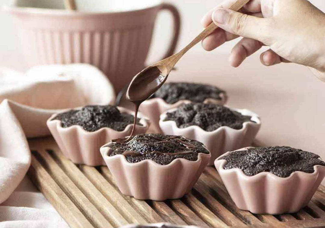 Eine Hand träufelt Schokoladensoße über frisch gebackene, flüssige Schokoladenkuchen in Keramikförmchen, neben einer Kaffeetasse.
