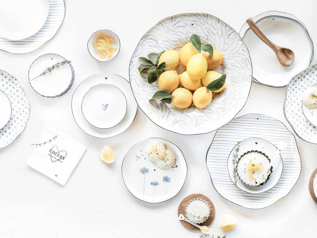 Elegante Tischdekoration mit verschiedenen Tellern mit Blumenmustern, einer Schale mit Zitronen als Tafelaufsatz und verstreuten Kerzen auf weißem Hintergrund.