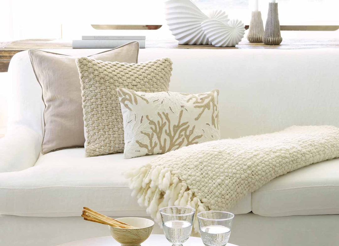 Ein weißes Sofa mit zwei Zierkissen und einer strukturierten Überwurfdecke, dazu ein Tablett mit einer kleinen Schüssel und Glaswaren auf einem Couchtisch.