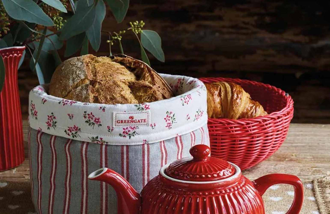 Eine rote Teekanne neben zwei Körben mit Brot, einer mit Blumenmuster und einer rot, auf einem Holztisch.