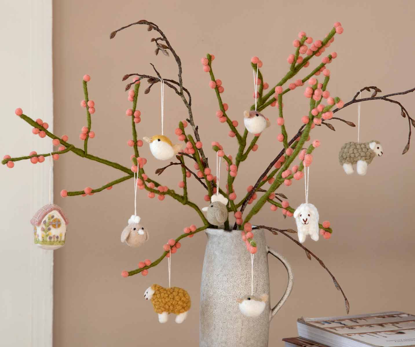 Ein dekoratives Arrangement aus Zweigen mit rosa Knospen und verschiedenen daran hängenden kleinen Tierfiguren, ausgestellt in einer grauen Vase auf einem Tisch.