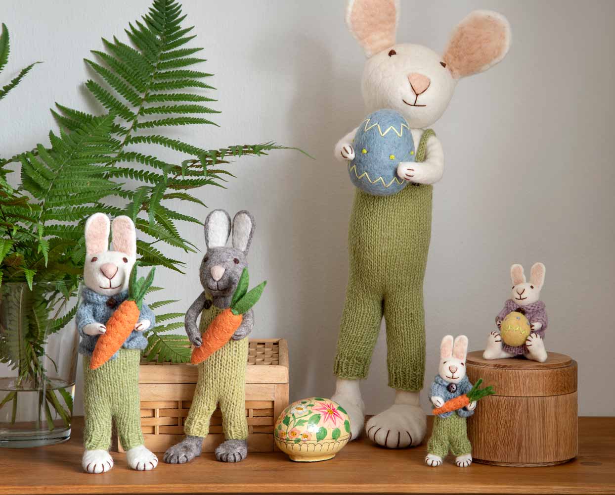Eine Sammlung von Kaninchenfiguren in grünen Overalls, einige mit Karotten und Ostereiern, ausgestellt auf einem Holztisch neben einer Farnpflanze.