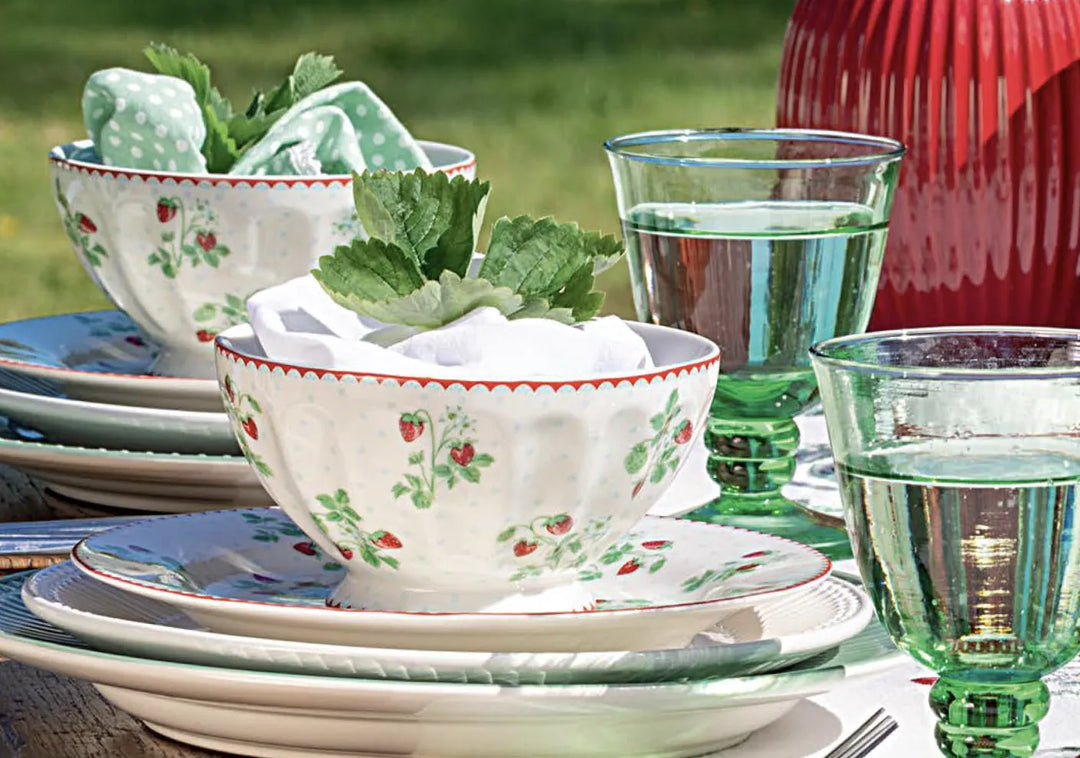 Zwei Gedecke mit Schalen und Tellern mit Erdbeermuster, grünen Glaspokalen, Servietten und Grünzeug auf einem Tisch im Freien.