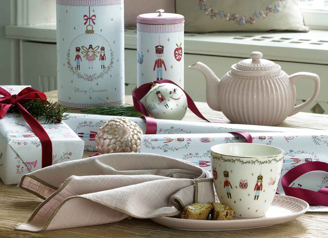 Tisch mit weihnachtlichem Teeservice aus Porzellan, bestehend aus Teekanne, Tasse und Zuckerdose, sowie verpackten Geschenken mit roten Bändern und Dekoelementen, arrangiert auf einer Holzoberfläche mit Blick auf ein Fenster.