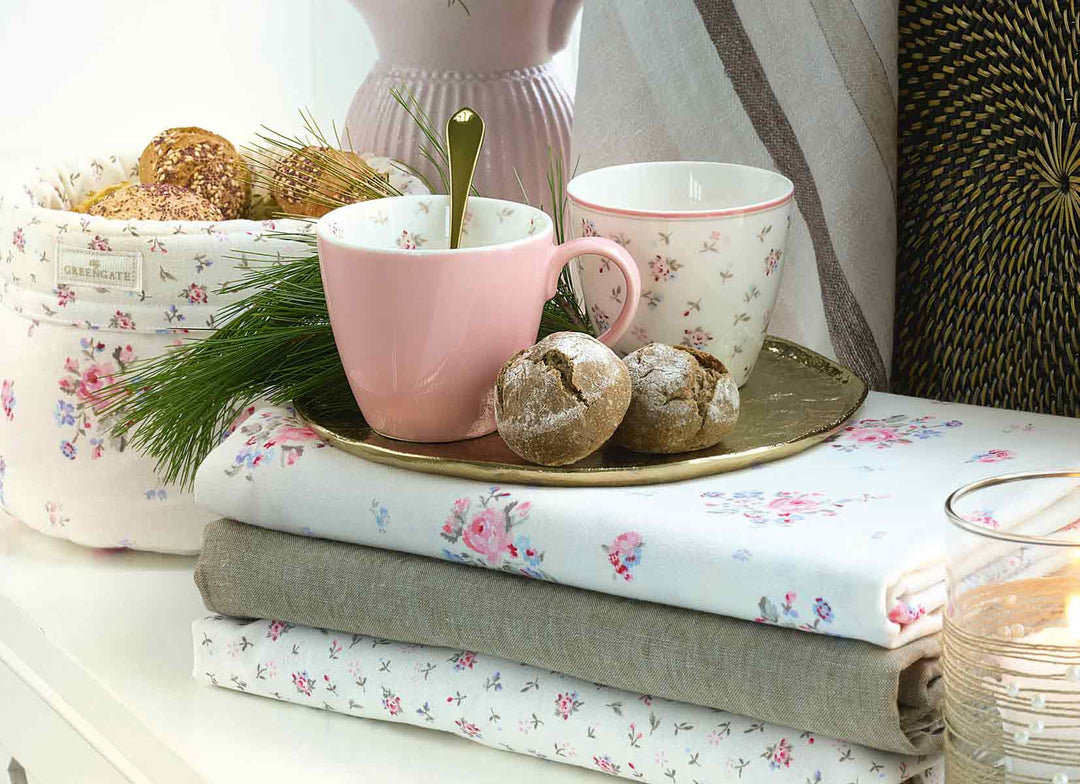 Eine rosa Tasse, eine Schüssel mit Blumenmuster und ein Teller mit Gebäck auf einem Stapel gefalteter Stoffe, neben einer Lampe und einer Pflanze auf einem Tisch.