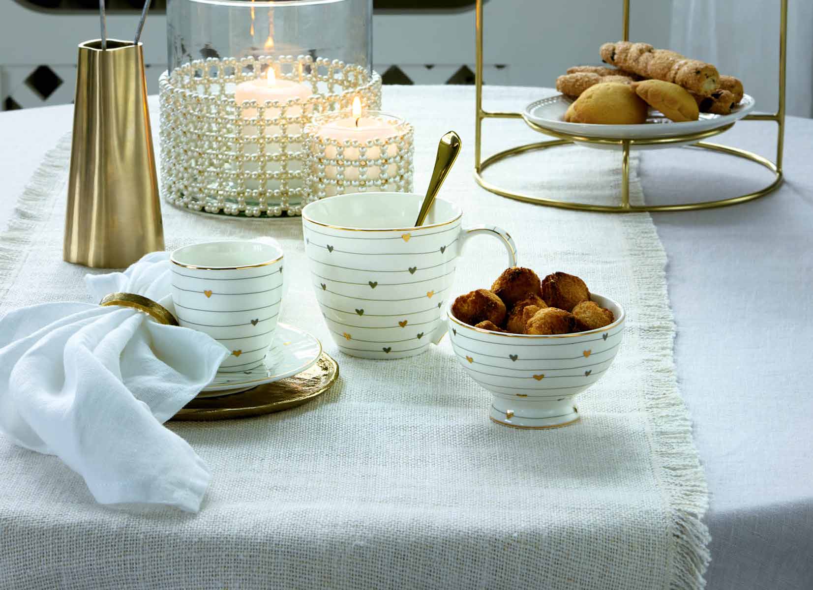 Auf einem mit einer weißen Tischdecke gedeckten Tisch stehen eine Teetasse mit Untertasse, eine Schüssel mit Gebäck, ein Becher, eine weiße Leinenserviette, eine goldfarbene Karaffe und eine Kerze in einem mit Kristallen verzierten Glashalter.