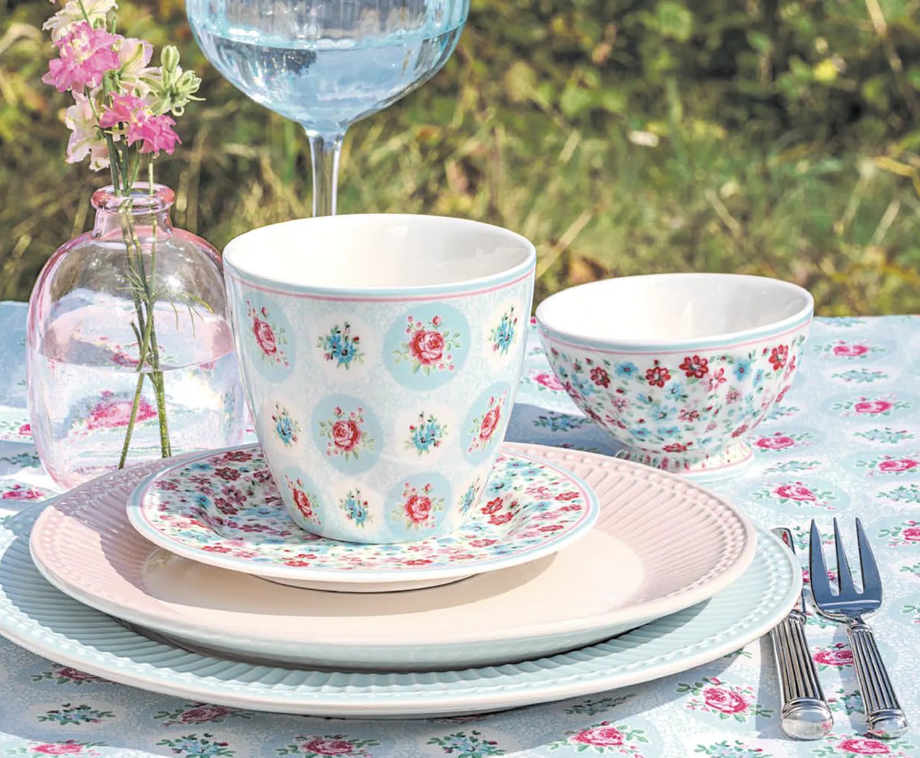 Ein Gedeck mit pastellfarbenem, blumengemustertem Geschirr, einer Tasse, einer Schale, Besteck, einem Weinglas und einer kleinen Vase mit rosa Blumen auf einer passenden Tischdecke.