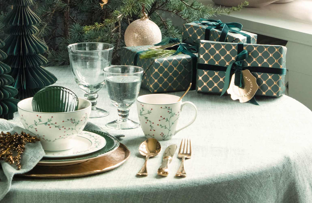 Eine festlich gedeckte Tafel mit Tellern, Tassen, Besteck und verpackten Geschenken auf einer hellgrünen Tischdecke, geschmückt mit Weihnachtsdekorationen.