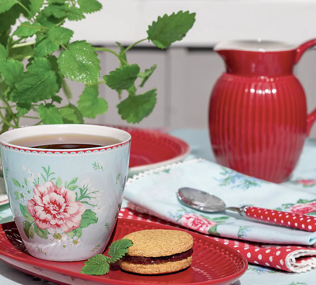Eine geblümte Teetasse mit Tee, ein mit Marmelade gefüllter Keks, ein roter Teller, eine rote Kanne, ein Löffel auf einer geblümten Serviette und eine grüne Pflanze auf einem Tisch.