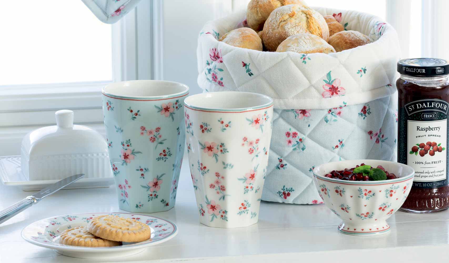 Ein Frühstückstisch mit zwei geblümten Tassen, einer dazu passenden Schale mit Beeren, einem Teller mit Keksen, einem Glas Himbeermarmelade, einer Butterdose und einem Korb mit Brötchen in einem Steppbezug.