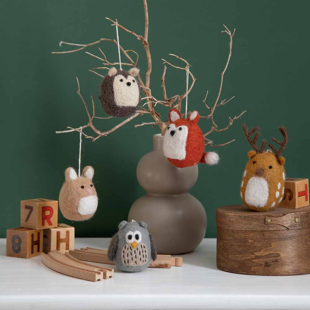 Eine Eule, ein Fuchs (Gry & Sif - Anhänger Fuchs Filz), ein Reh und ein Hase aus Filz hängen an einem minimalistischen Astständer in einer Vase, auf einem Tisch stehen Holzklötze und Eisenbahnschienen