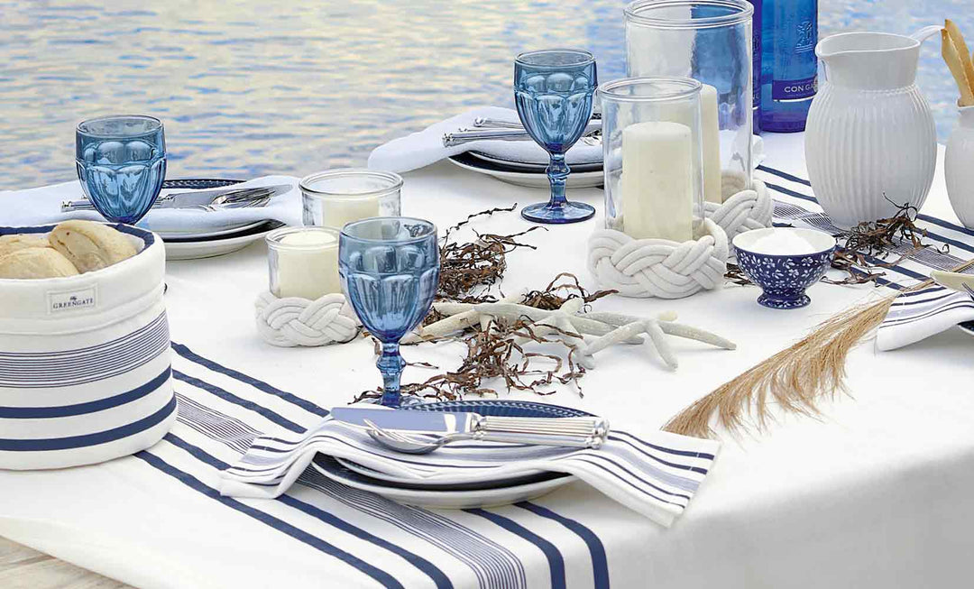 Eleganter Esstisch mit blauem Glasgeschirr, gestreifter Tischwäsche, weißem Geschirr und nautischer Dekoration, am Wasser arrangiert.